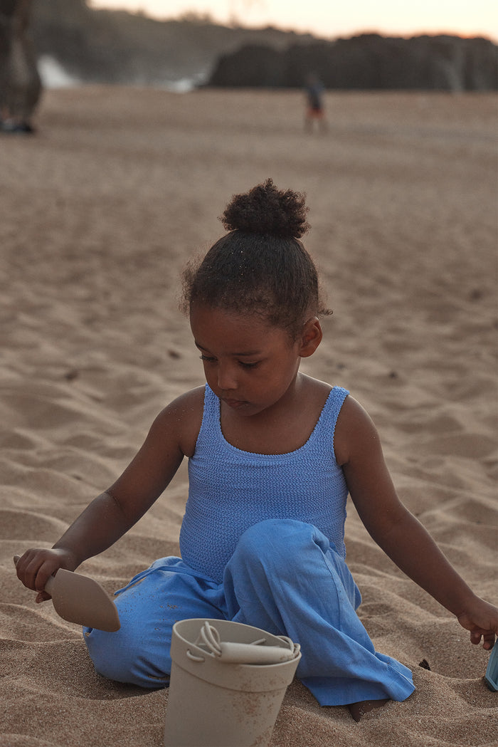 Front view of Tuscany Pant Kids in Pool blue showcasing relaxed wide-leg knit pants with child model seated on a sandy beach