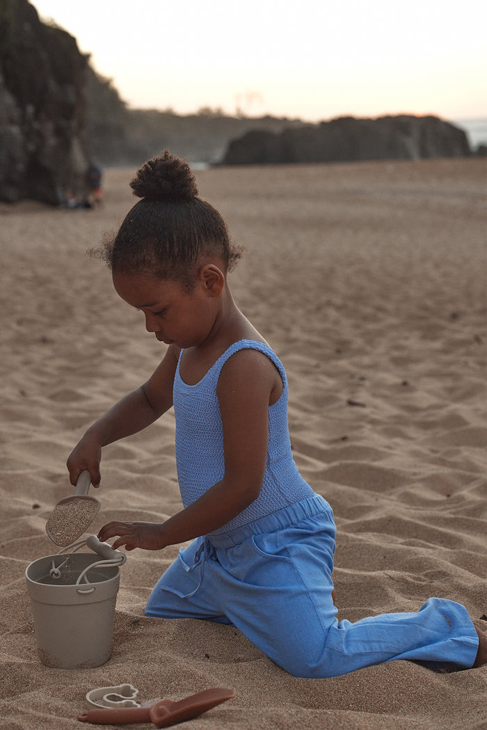 Side view of Tuscany Pant Kids in Pool blue on a child, showing relaxed fit and drawstring waist on a sandy beach