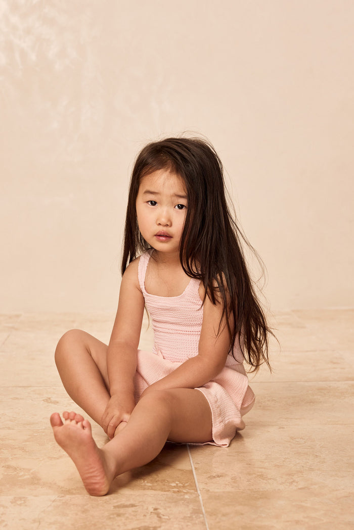 Seated front view of Sardinia Kids one-piece in Powder Pink Crinkle showing scoop neckline on beige background