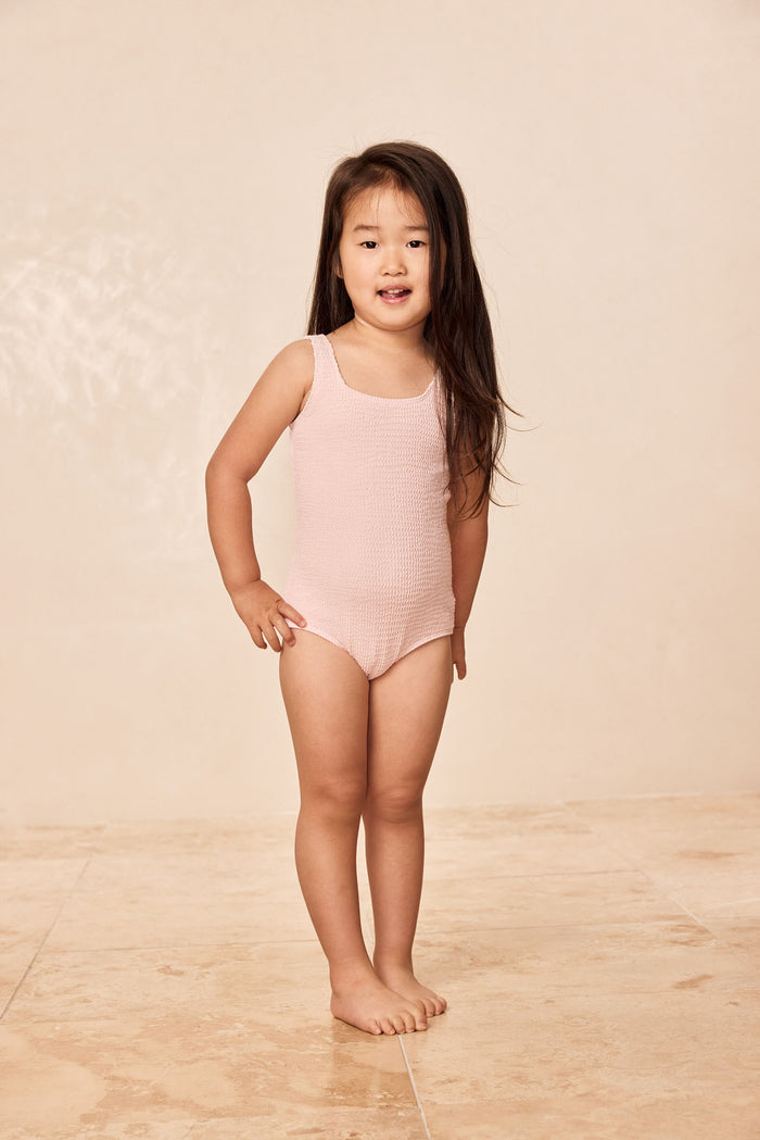 Front of Sardinia One Piece Kids, Powder Pink Crinkle, scoop neckline, regular fit on child model, beige background