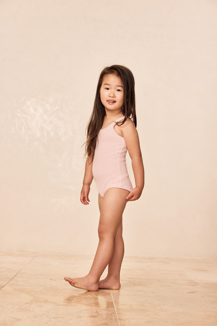 Side view of Sardinia One Piece Kids in Powder Pink Crinkle showing scoop neck and textured crinkle fabric on beige background