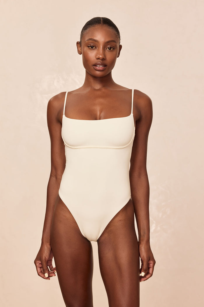 Front view of Sausalito One Piece in Ivory showcasing scoop neckline, under bust support, thin adjustable straps and high cut leg on beige background