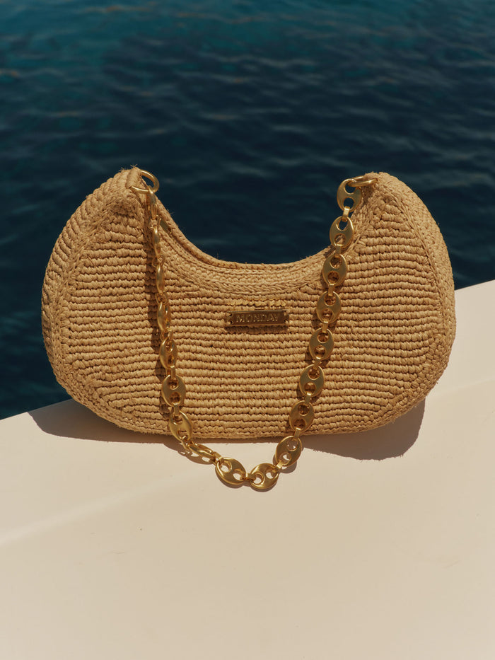 Front view of Siracusa bag in Natural raffia showcasing gold metal chain strap and gold MONDAY logo plate against a deep-blue sea background