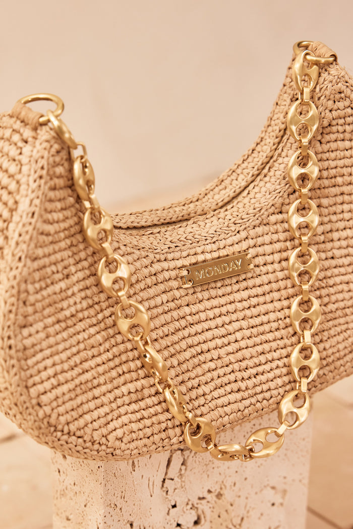 Angled close-up of Siracusa bag in natural raffia showcasing gold chain strap and MONDAY logo with neutral stone pedestal background