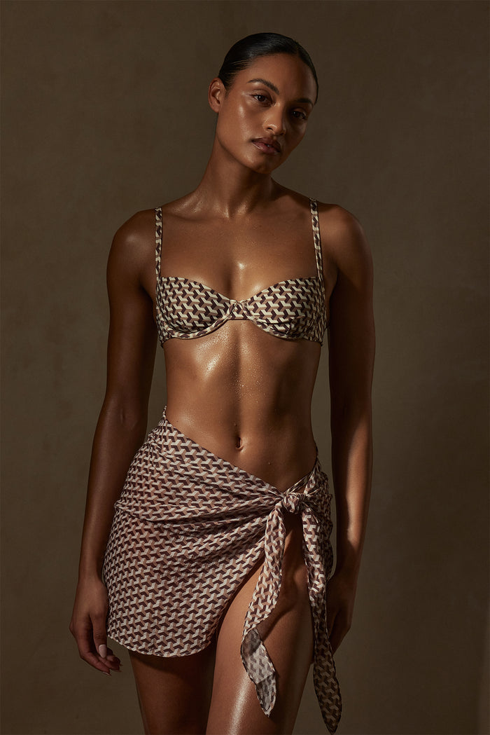 Model wearing brown geometric-print balconette bikini top and matching sarong, glistening skin, warm brown background.