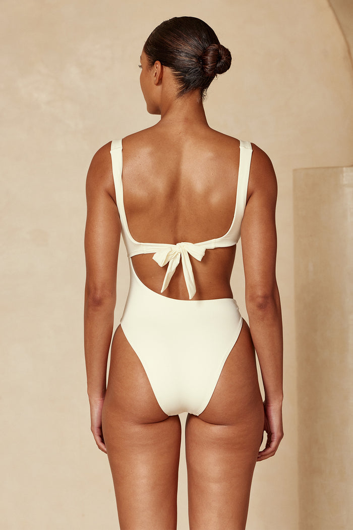 Back view of woman wearing high-cut cream one-piece swimsuit with bow tied at mid-back, hair in a low bun