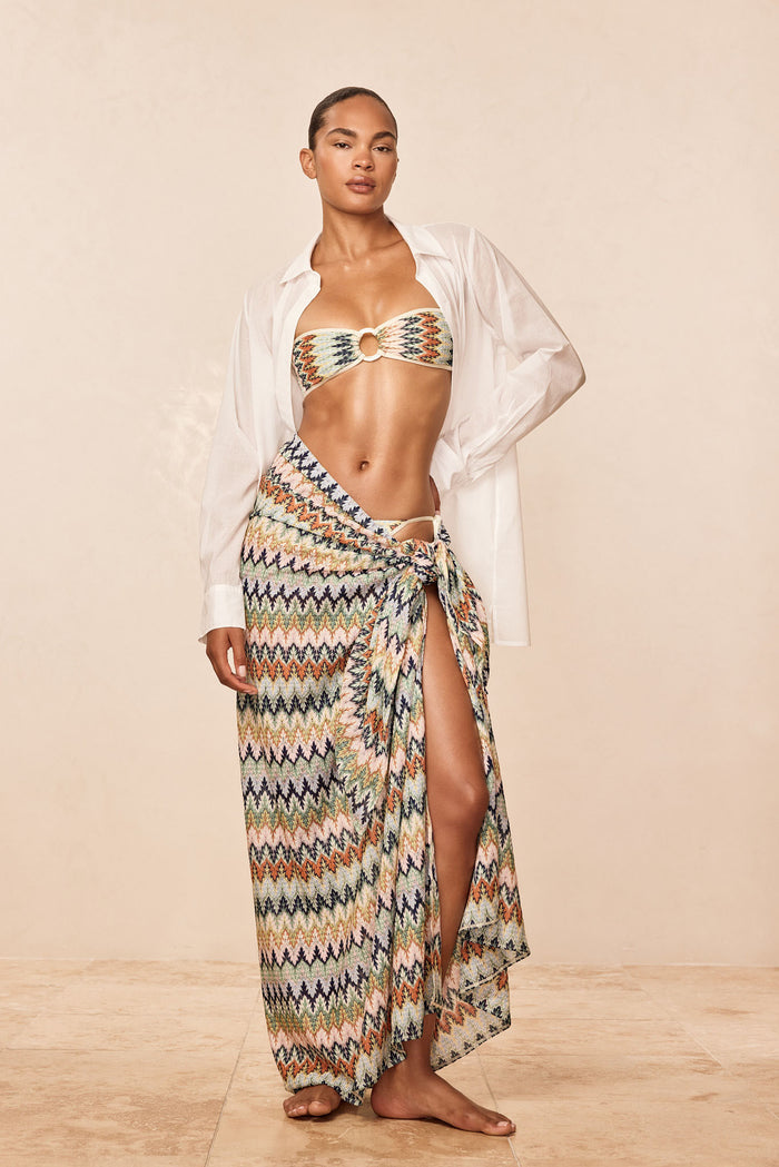 Three-quarter view of St Tropez Sarong Porto Cervo Multi showcasing multicolor zigzag pattern, high side slit, and tie detail on a model with a beige studio background