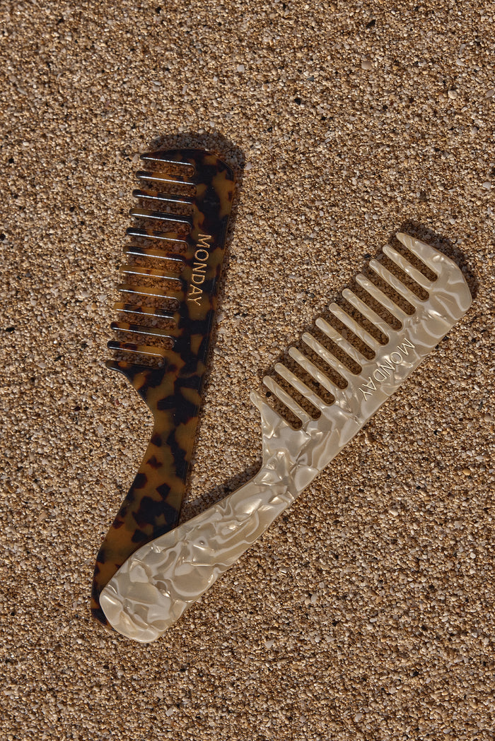 Two wide-tooth combs on sand: tortoiseshell and pearlescent styles, each embossed with the word MONDAY.