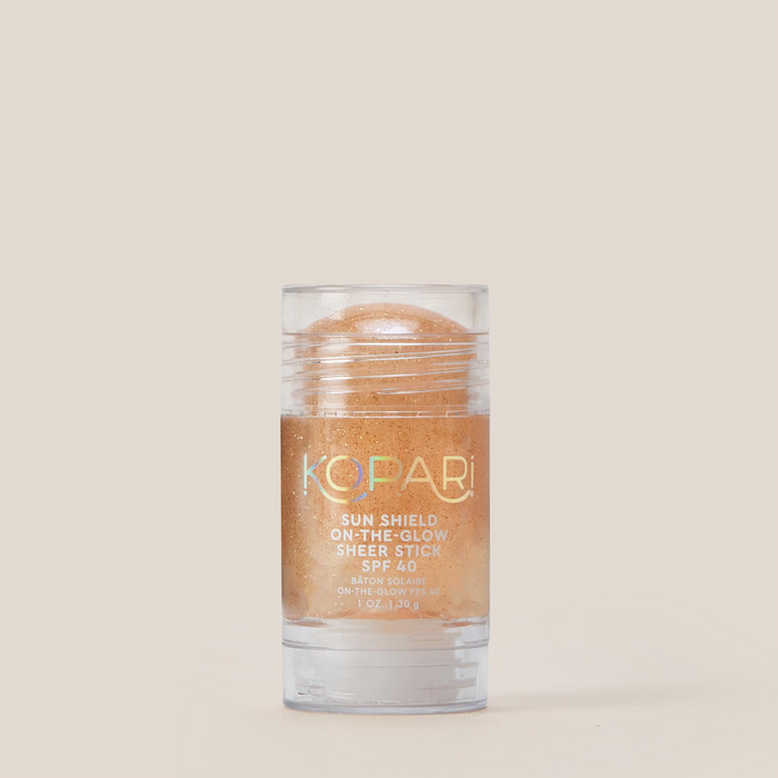 Front view of Kopari Sun Shield On-The-Glow Sheer Stick SPF 40 showing sheer golden glow stick in clear tube on beige background