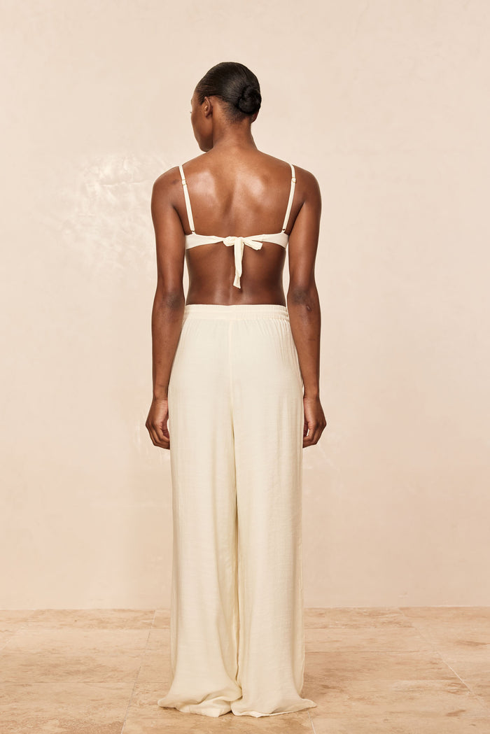 Back view of Tahiti Pant in ivory on model showing high-waist, wide-leg fit with tie-back bikini top on beige background