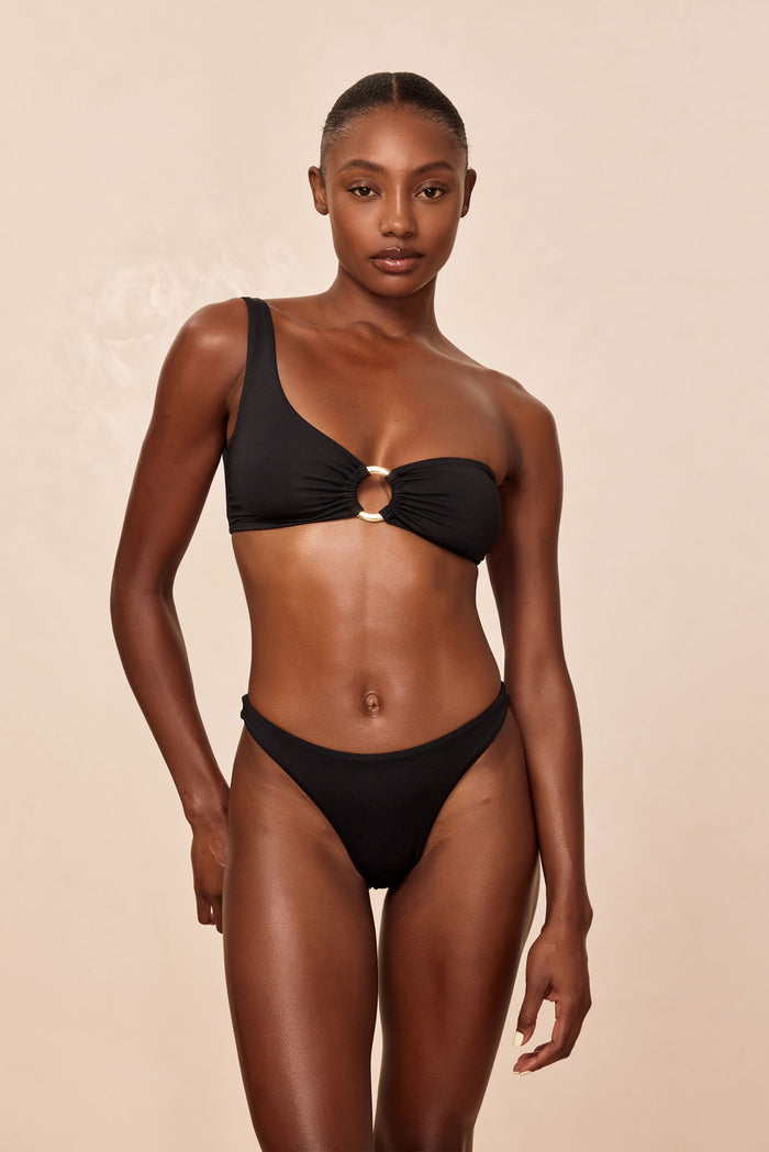 Front view of Tamarama Bottom in black on model showcasing cheeky coverage, thin sides, and high leg cut with neutral beige background