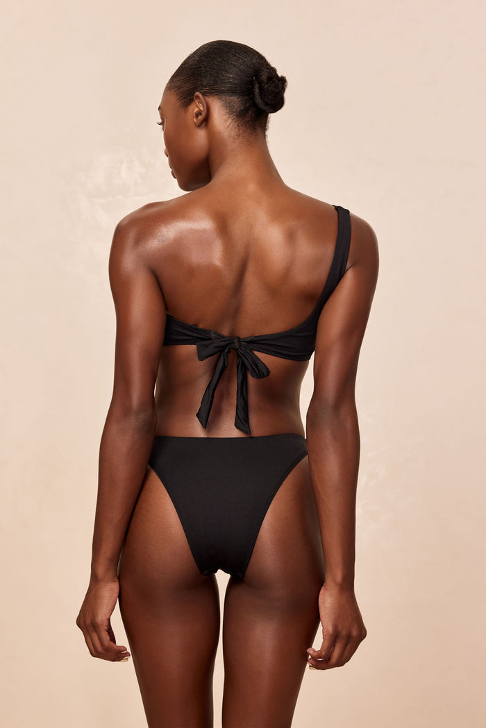 Back view of Tamarama Bottom in black showcasing high leg cut, thin sides and cheeky coverage on model against beige background