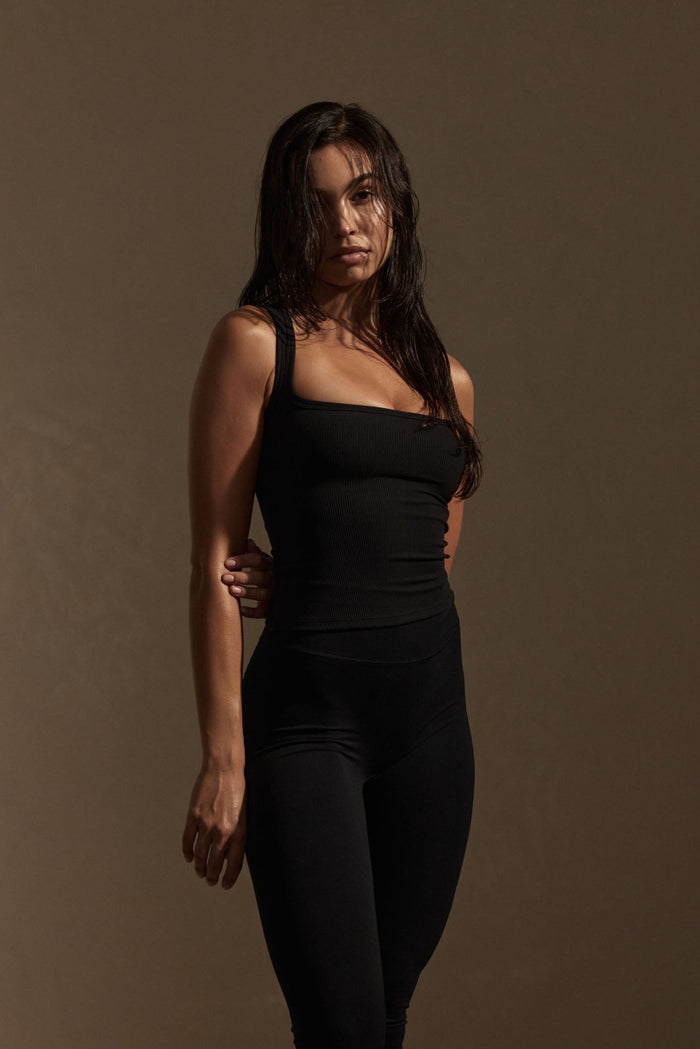 Three-quarter view of Tash Tank Long in Black Rib showing thin rib texture and wide square neckline, fitted on model against a brown studio background