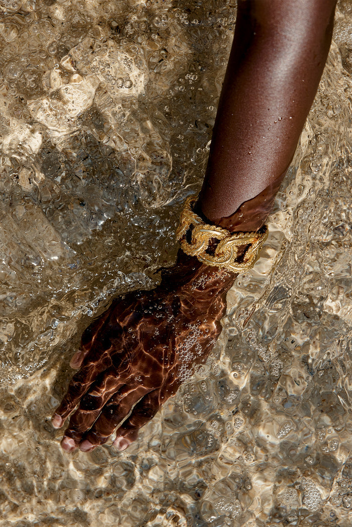 Dark-skinned forearm and hand submerged in rippling shallow water, wearing a chunky braided gold bracelet.