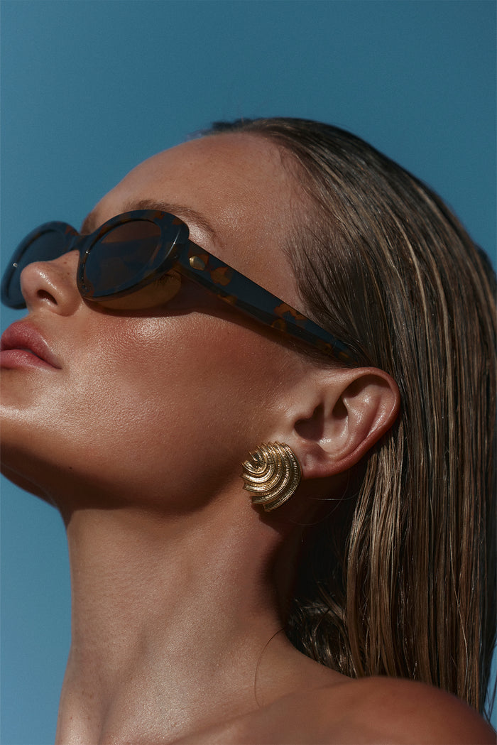 Close-up profile of Trancoso Earrings in gold showcasing layered ribbed shell design on model against blue sky