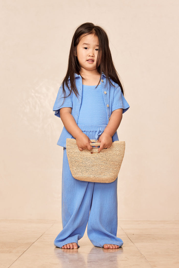 Front full-length of Tuscany Pant Kids in Azure Crinkle showcasing smocked waistband, flare leg, crinkle texture on beige background