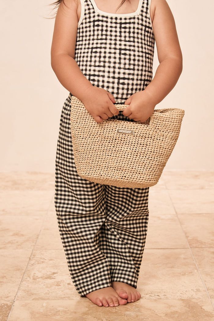 Front view of Tuscany Pant Kids in Black Gingham showcasing smocked waistband and flare leg on a child holding a straw tote on beige tile