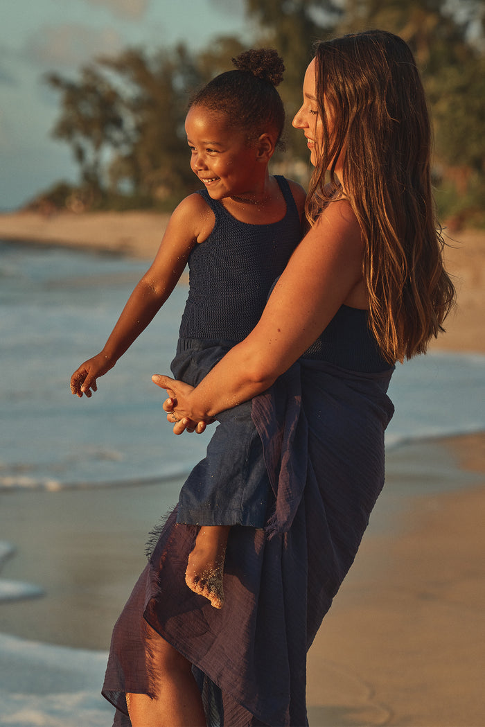 Side view of Tuscany Pant Kids in Midnight worn by a child, showcasing cropped mid-rise fit and navy fabric on a sandy sunset beach