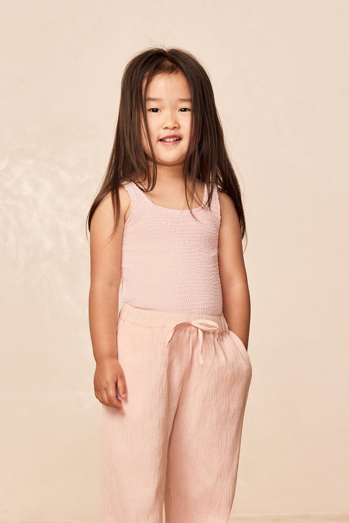 Front of Tuscany Pant Kids in Powder Pink Crinkle showing smocked waistband and wide flare leg on child, pale beige backdrop
