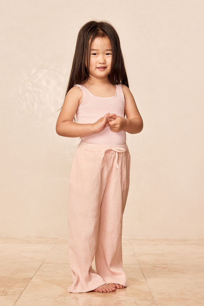 Front view of Tuscany Pant Kids in powder pink crinkle with smocked waistband and flared legs on child, beige background