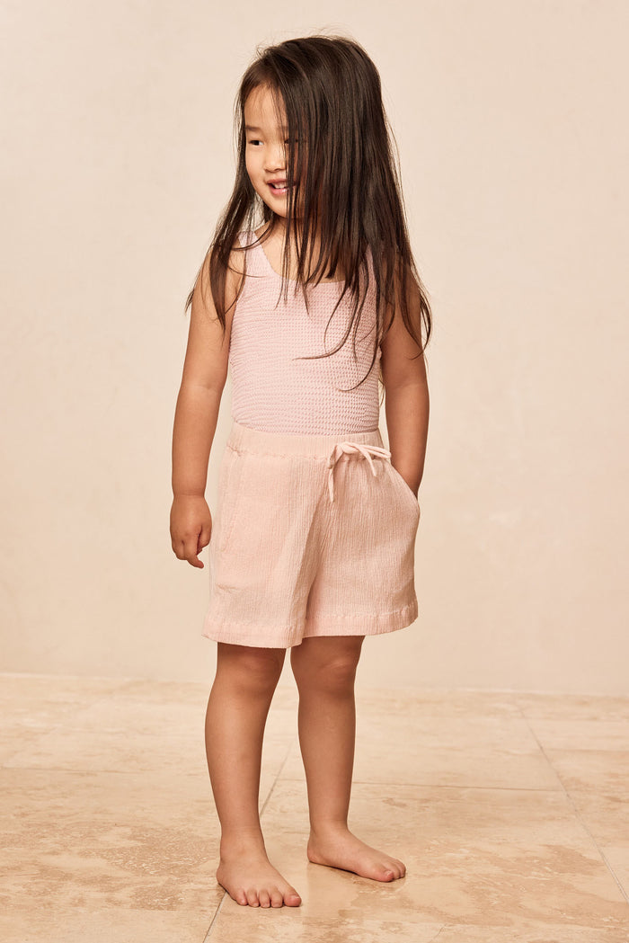 Three-quarter view of Tuscany Short Kids in powder pink shorts showing elastic waist with faux tie and pockets on neutral background