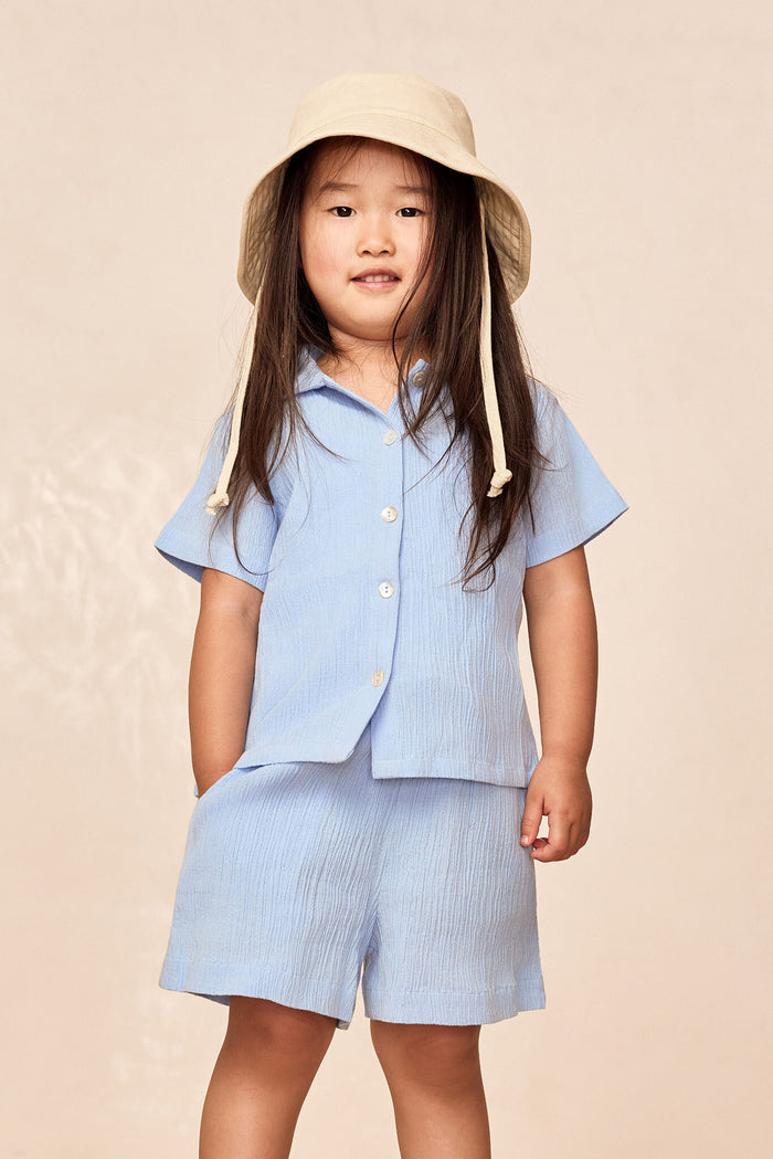 Front of Tuscany Kids shorts in Sky showing crinkled fabric, elastic waist and faux tie on child model, beige background