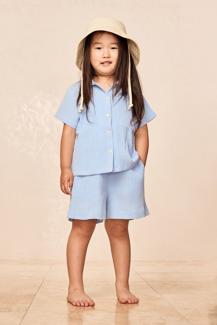 Front view of Tuscany Shorts Kids in Sky showing elastic-waist pull-on shorts and matching button-front top on child model against beige background