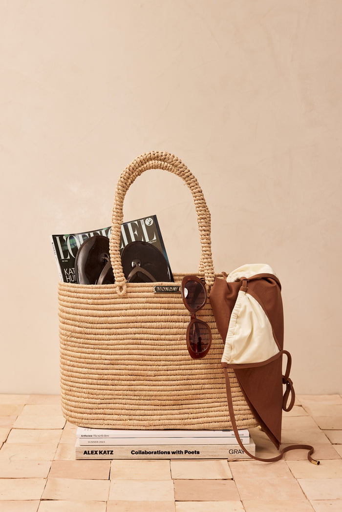 Front view of Valencia Bag - Large, woven raffia tote with braided handles and gold MONDAY plaque on beige backdrop