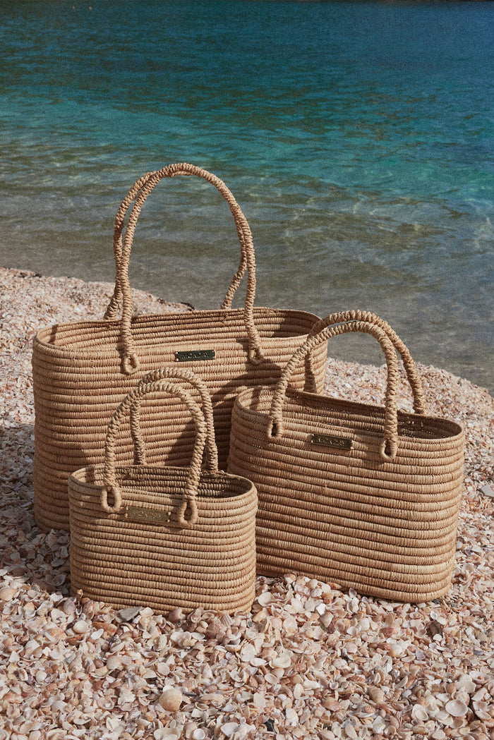 Angled view of Valencia mini woven straw bag with rope handles, paired with larger sizes on a shell beach with blue water