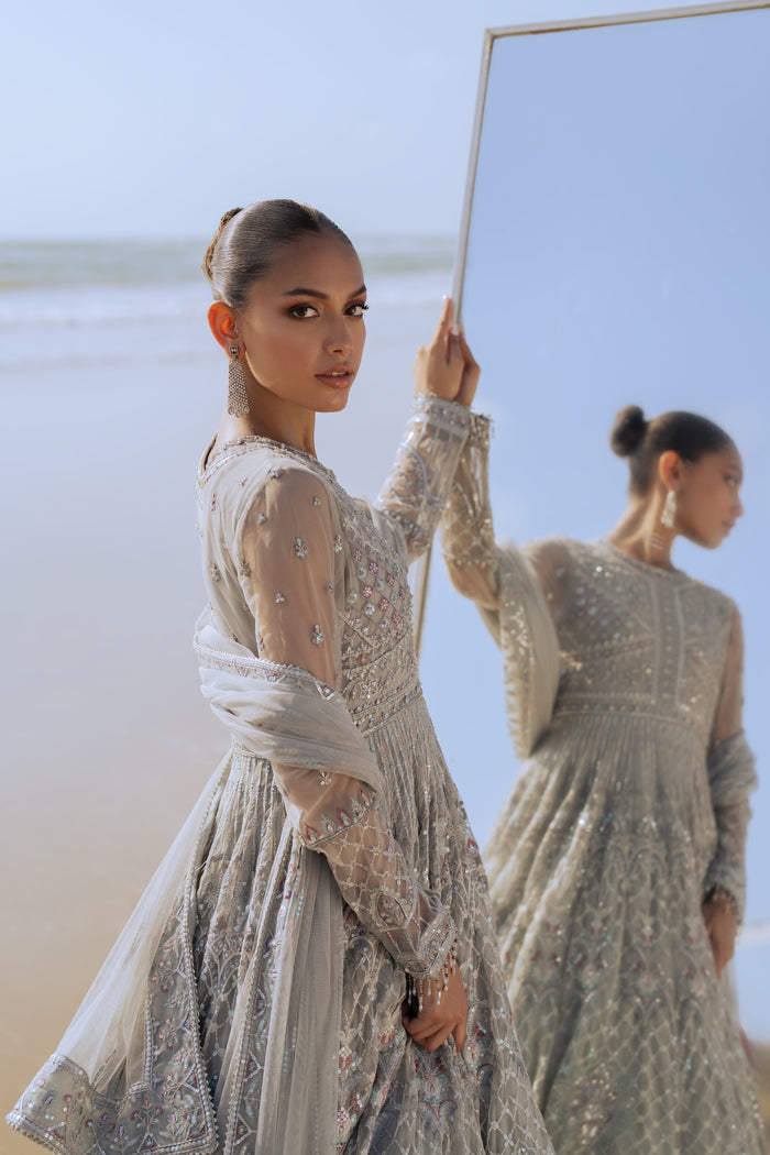 Model wearing full NAQSHI Valeria 3PC: expertly embellished silver net frock with hand‑adda detailing, plain PK raw‑silk pants & matching embroidered dupatta for a refined formal look.