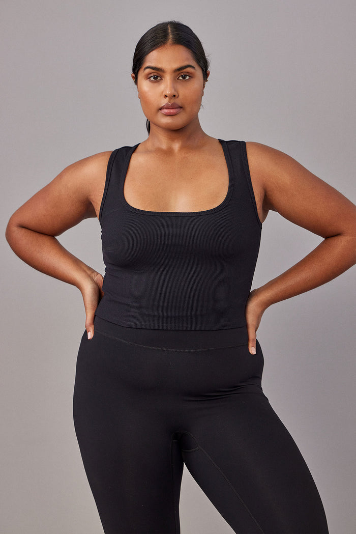 Woman wearing black ribbed square-neck tank and high-waist black leggings, hands on hips, gray background