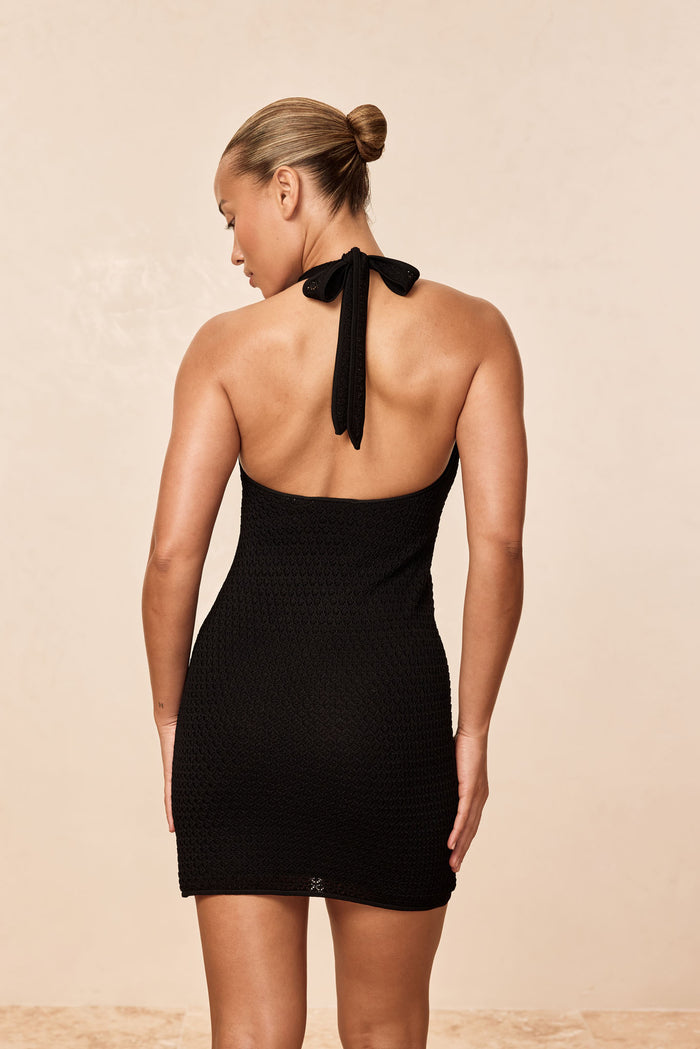 Back view of Waikiki Mini Dress in black lace crochet showcasing halter tie, open back, and textured crochet fabric with neutral beige studio background