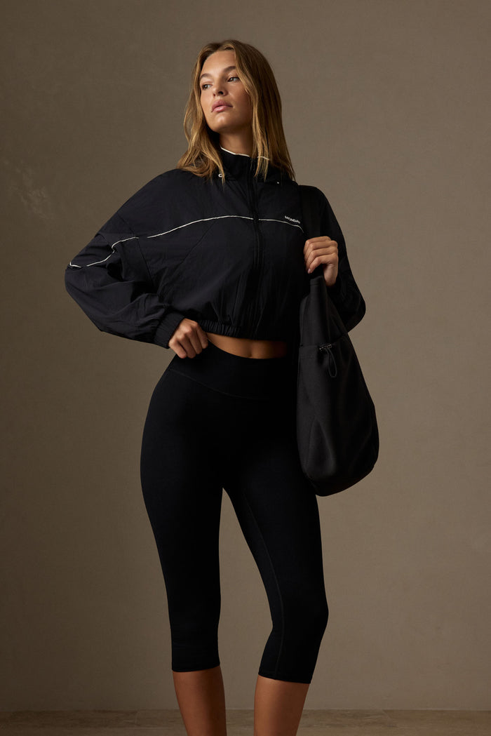 Female model wearing black cropped Westmount jacket with white piping and high collar, holding a black tote against a neutral background