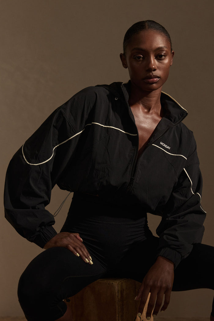 Cropped black windbreaker with white piping and MONDAY chest logo, worn by seated model