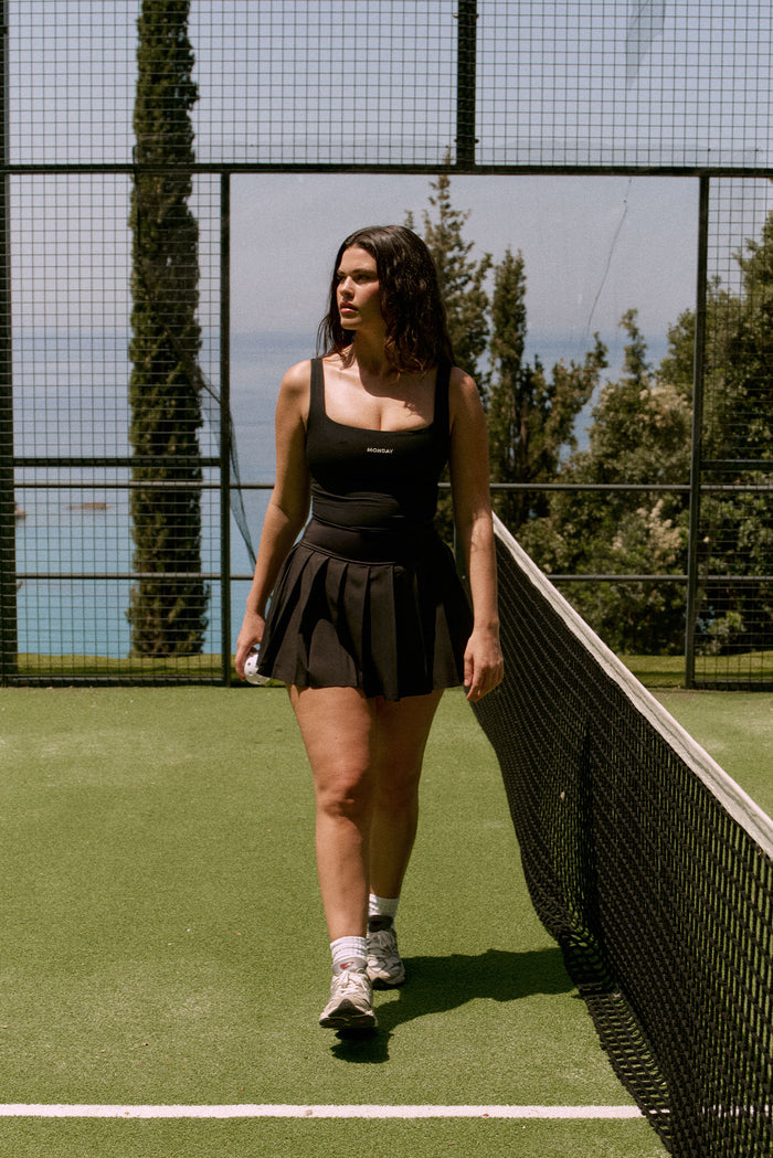 Front view of Wimbledon Skirt in black showcasing pleated lightweight skirt and fitted waistband with model on a tennis court