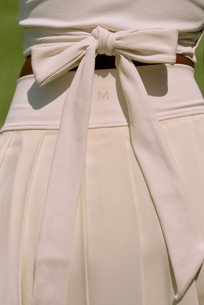 Close-up back of Wimbledon Skirt in ivory showing ribbed bow-tied waistband and centered M logo, tennis court background