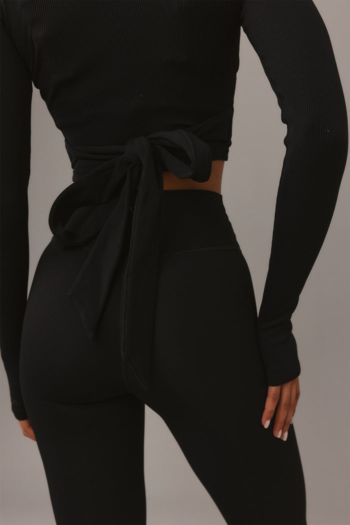 Back view of Yosemite Top in Black Rib showcasing ribbed texture, tied wrap waist, and long sleeves on gray background