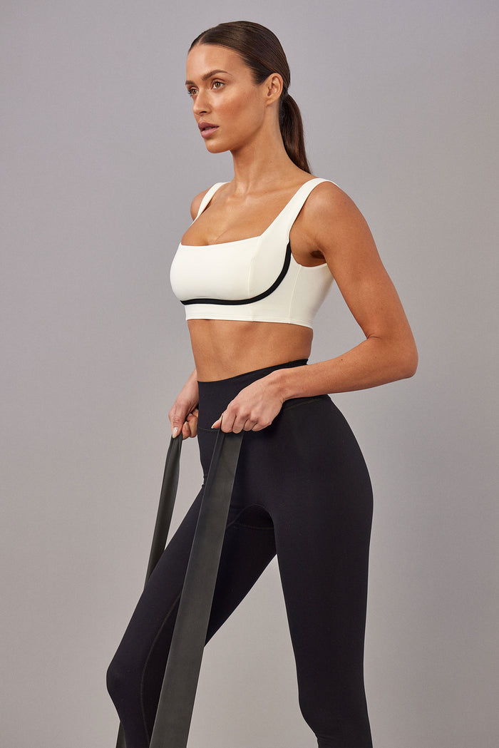 Three-quarter view of Zion Bra in ivory with black trim showing square neckline, wide straps, and supportive underbust band on model against gray background
