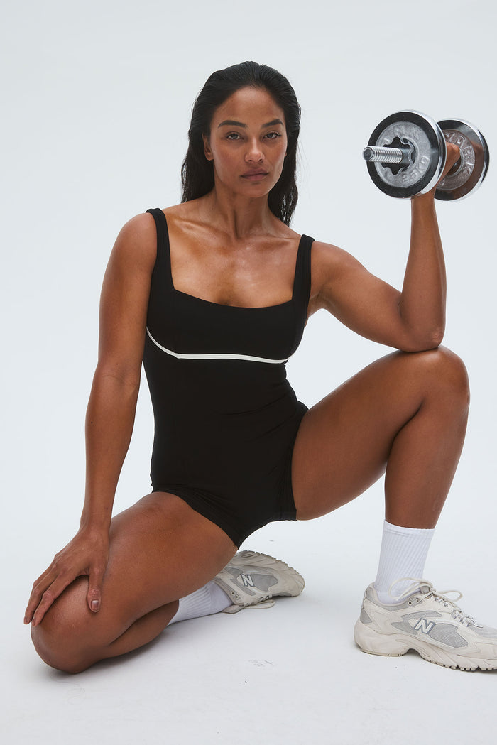 Front view of Zion Romper in black with ivory piping, fitted square-neck romper on model kneeling against white background