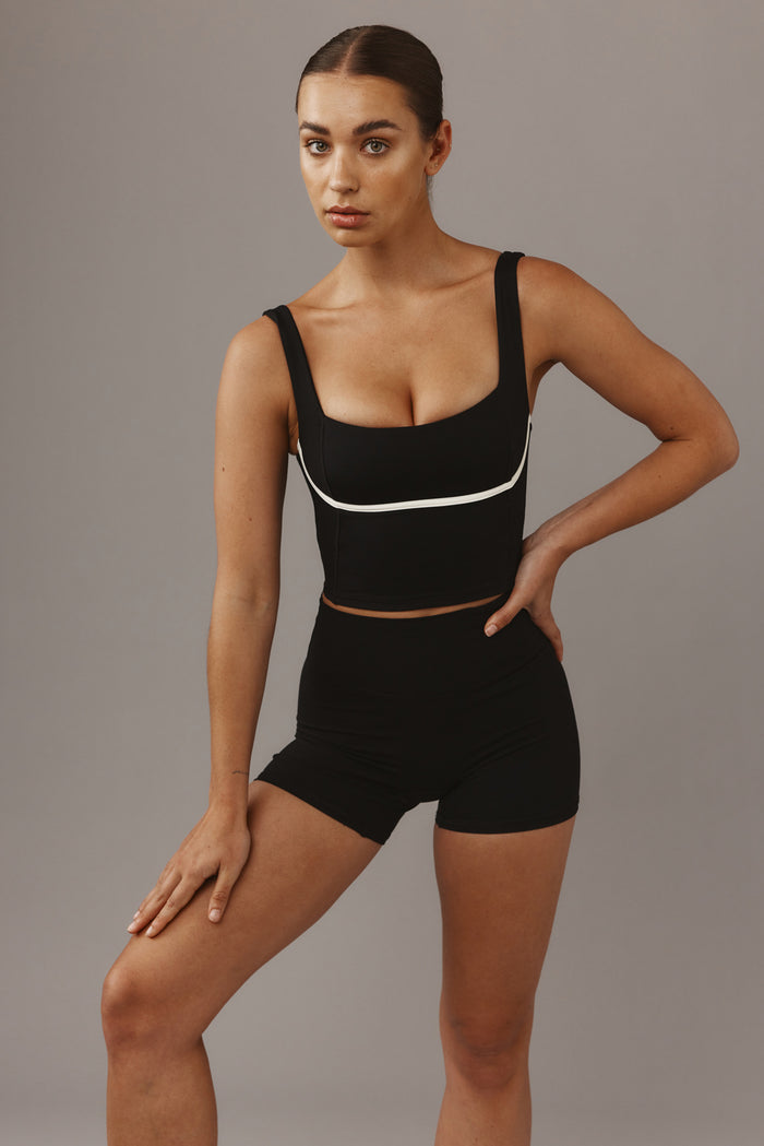 Woman wearing black cropped tank top with white piping and high-waisted black shorts, hand on hip against gray backdrop