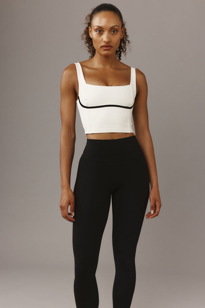 Model wearing white square-neck crop top with black piping and high-waist black leggings on gray background