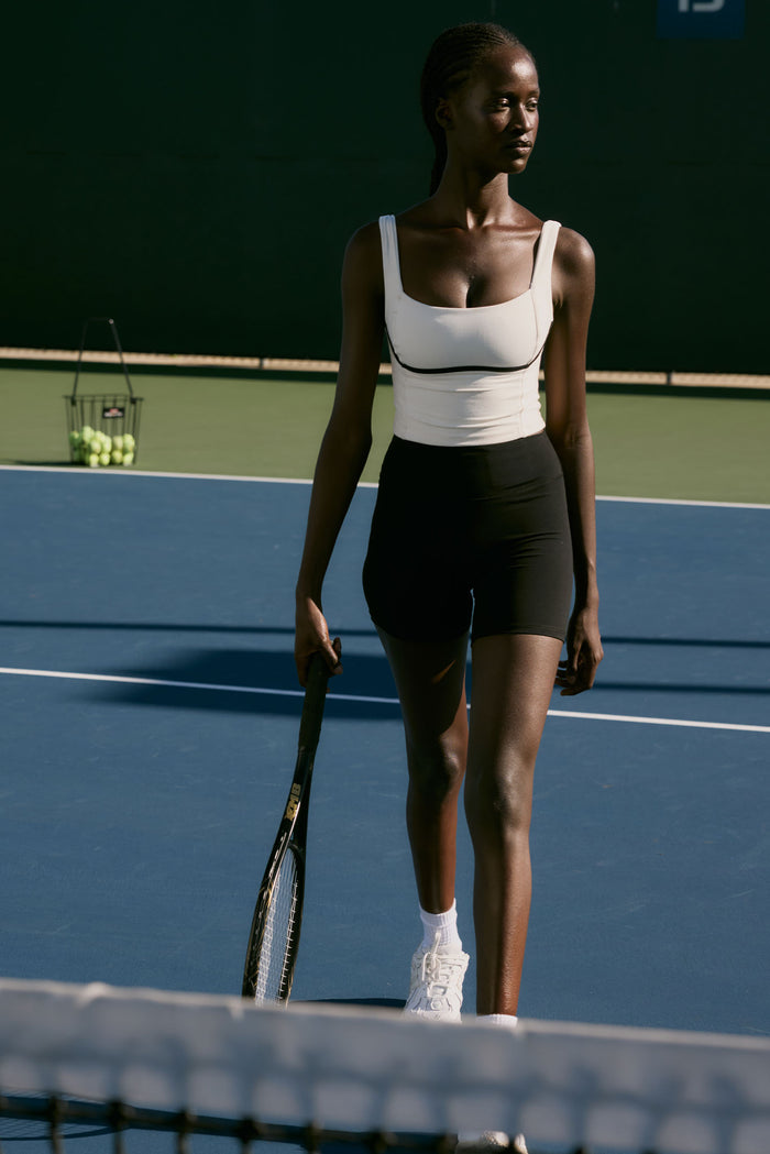 Woman on tennis court holding racquet, wearing white tank top and black shorts; basket of tennis balls at baseline.